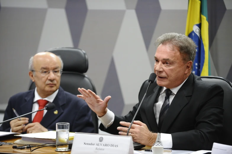 two men sitting at a table while one person holds his hands out