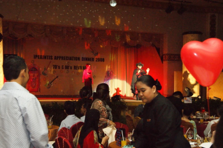 some people are sitting at tables and a man in the background is holding red heart balloons