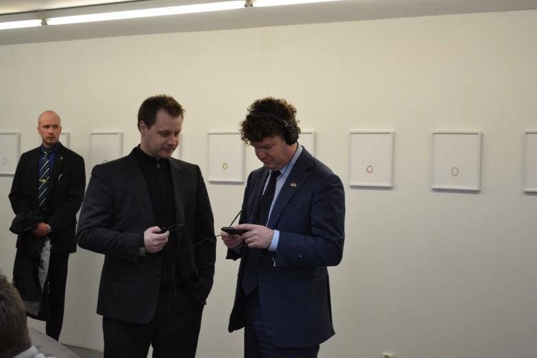 two men in suits and tie stand looking at soing on a cell phone