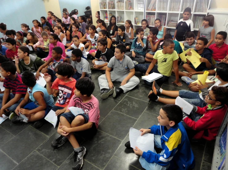 children in a classroom with adults and one boy sitting on the floor reading