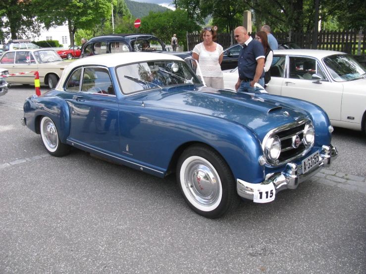 an old blue car sitting next to a building with people standing around it