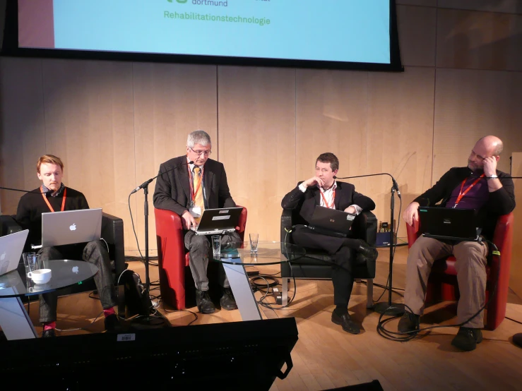 four men sitting in chairs around a laptop