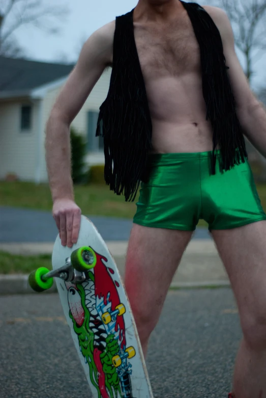 shirtless man wearing green trunks and fringed vest holding a skateboard
