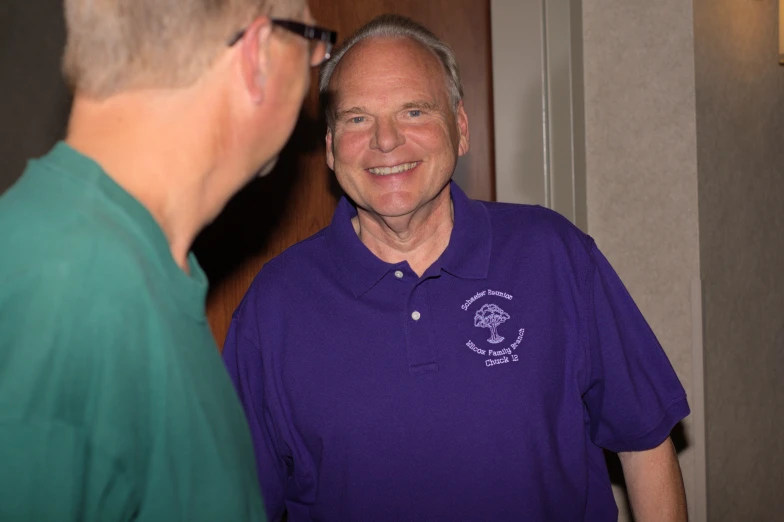 an older man wearing a purple shirt smiles as he talks to a younger man