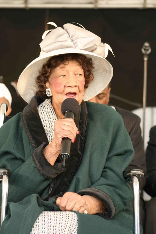 woman wearing a big white hat and green robe talking into a microphone