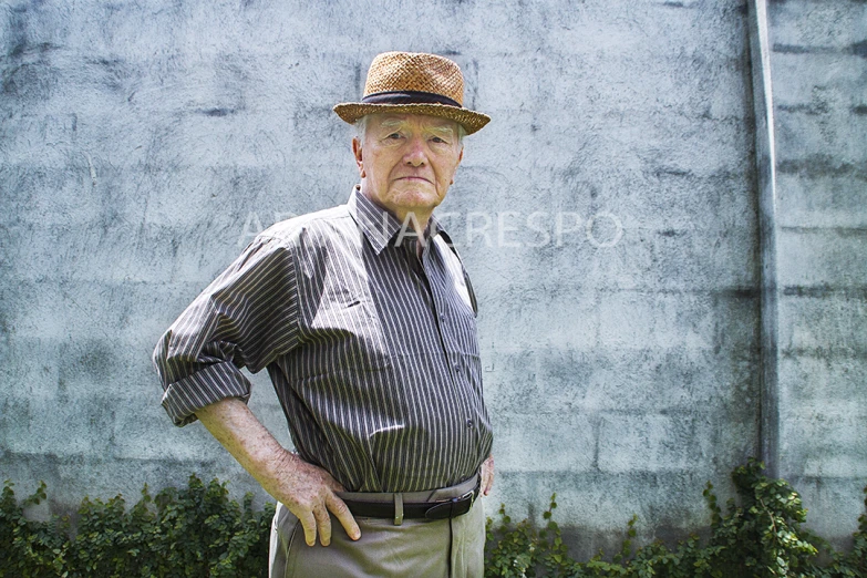 an old man wearing a hat stands against a cement wall