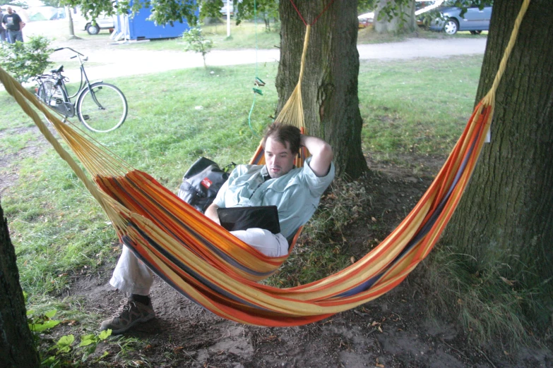 a man in an orange and yellow hammock reading a book while sitting next to a tree