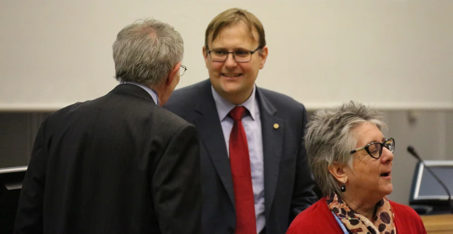 two older people in suits talk with a man in red tie