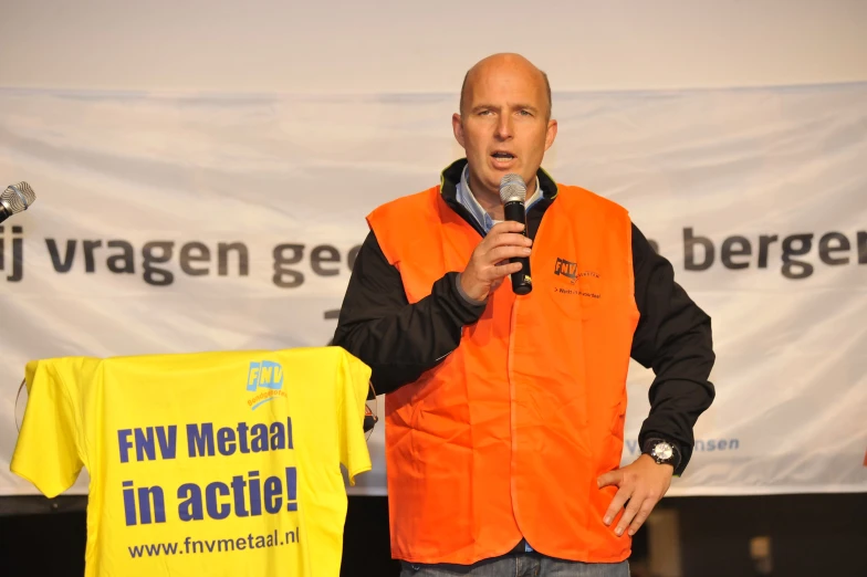 a man wearing an orange vest is standing in front of a sign holding a microphone