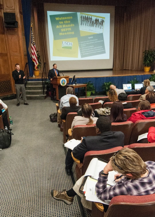 a man speaking at a podium to people seated in a classroom