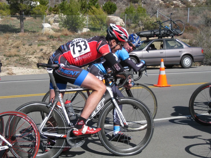 two people in bibs sitting on bicycles by a car