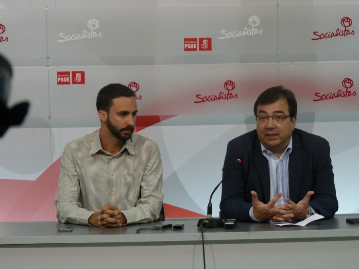 two men sit in front of microphones at a press conference