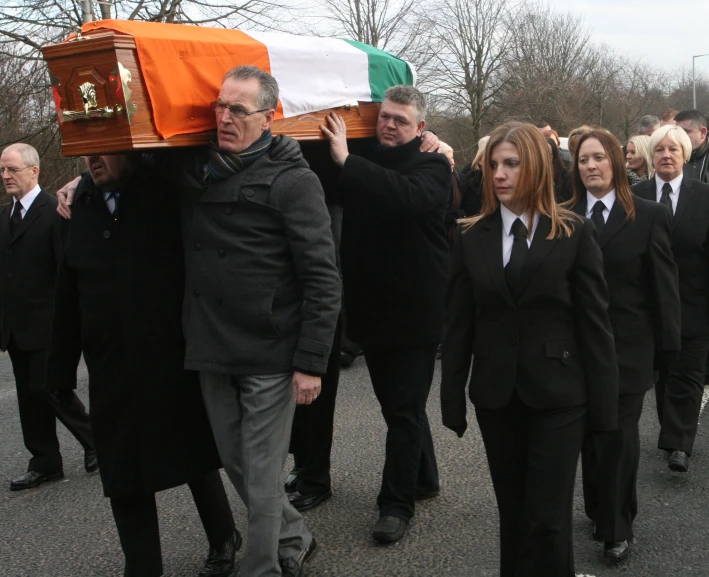 several men and women walking down a street carrying a casket