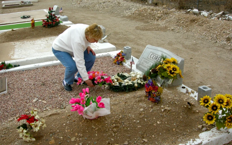 a woman kneels on a cemetery bench surrounded by flowers