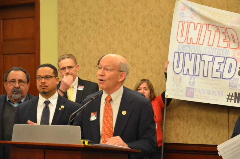 man in suit speaking at podium with sign next to him