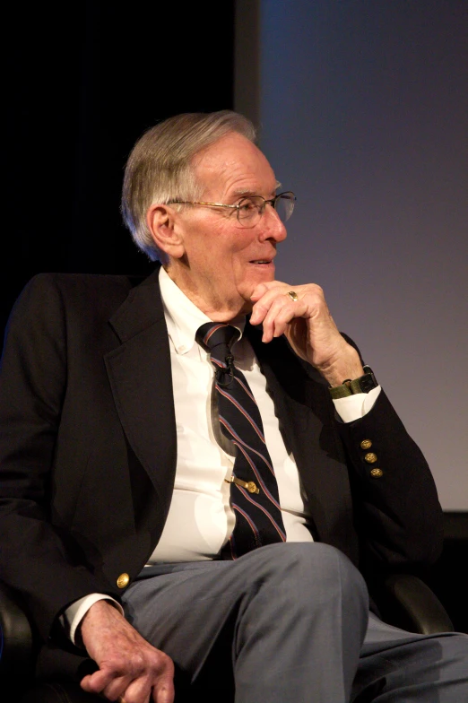 man in black jacket with dark tie seated with hands on his chin