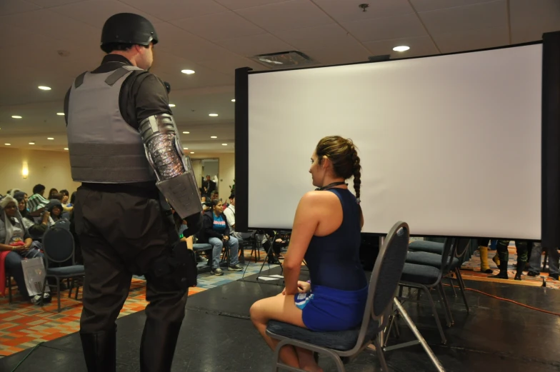 the woman is sitting in a chair and a man is in the back ground with his helmet on