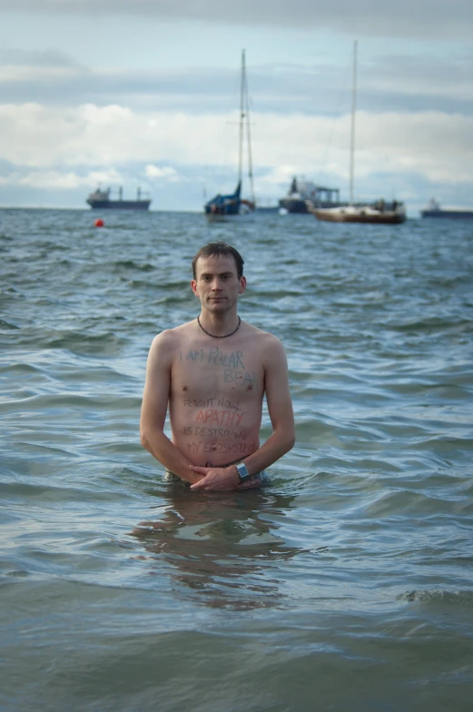 man stands shirtless in open water with sailboats in the background