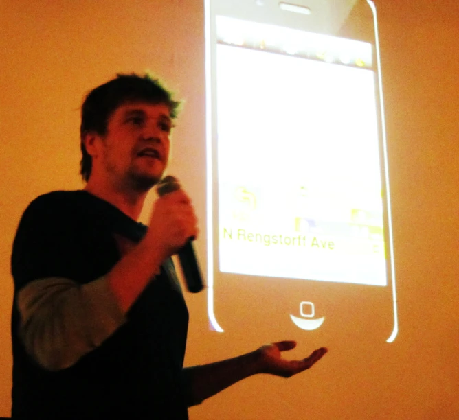 a man with a microphone stands at the front of a room and speaks to people in front of him a big screen in the shape of a phone