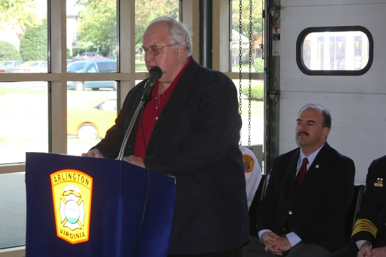 the man stands at a podium speaking while others sit on the porch