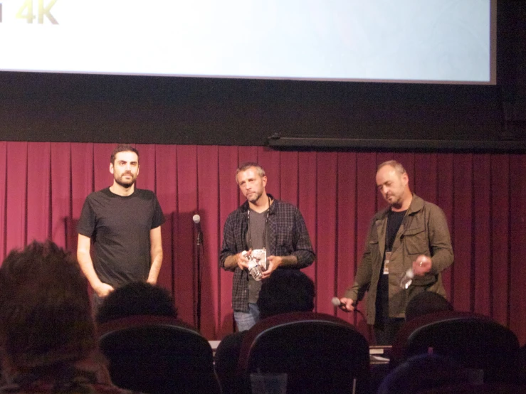 three men stand on stage while two microphones are hanging above them