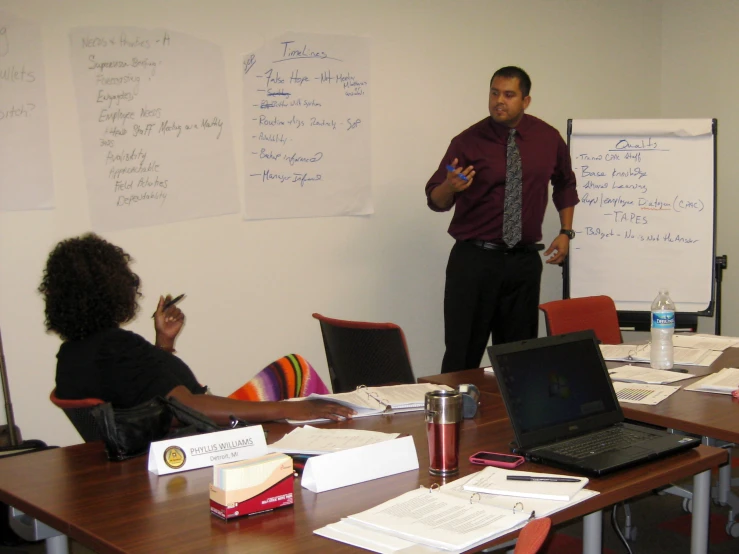 two woman and one man are sitting at tables in front of whiteboards with notes on it