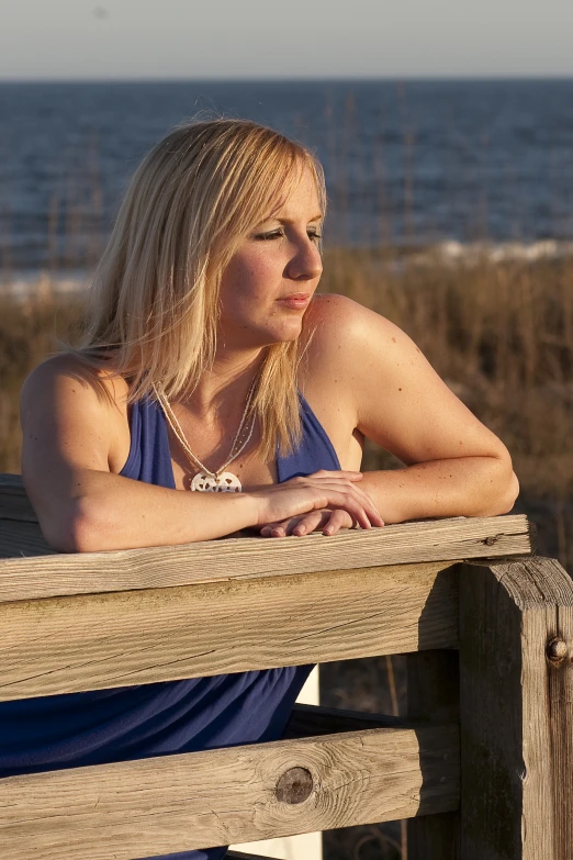 a beautiful blonde woman in a blue top sitting on a bench near the ocean