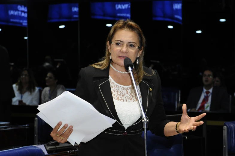 a woman in a black jacket holding a microphone and pointing at a paper