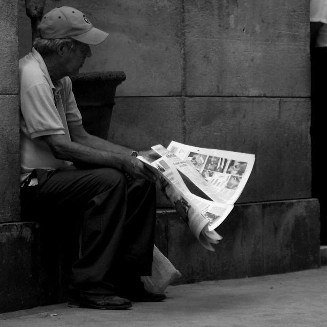 an old man sits on the sidewalk as he reads the newspaper