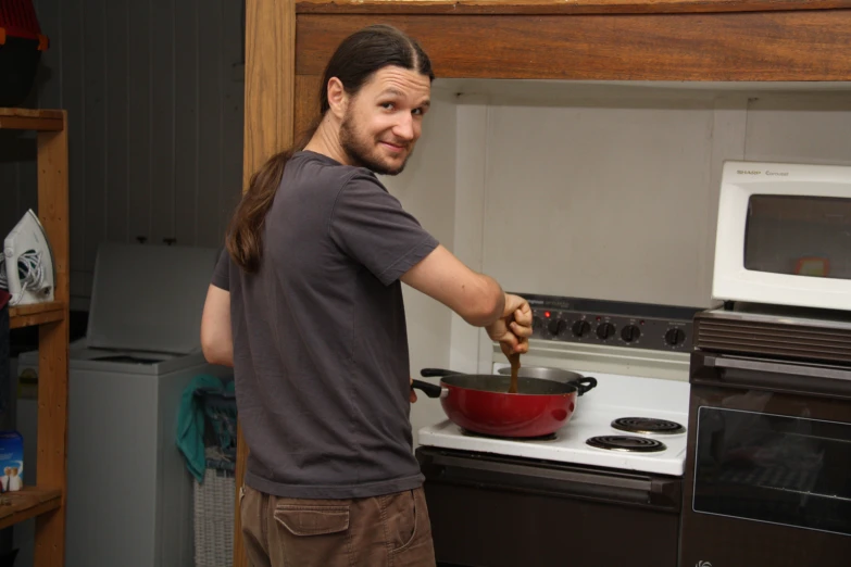 a man wearing a brown shirt and standing in front of an oven holding a red pan