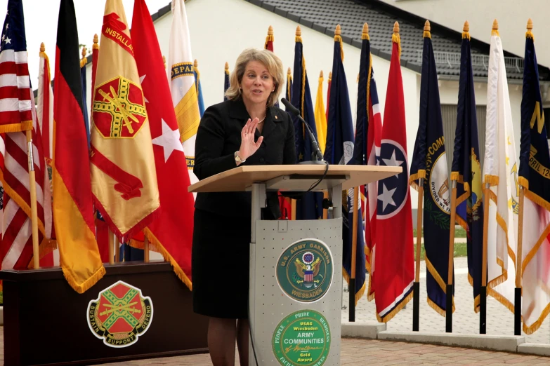 an older woman speaking from a podium in front of a collection of flags