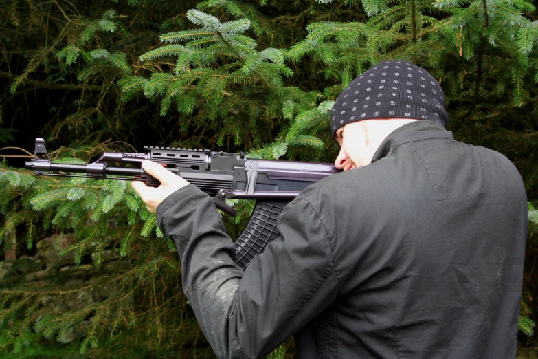 man with a gun and head covering by pine trees