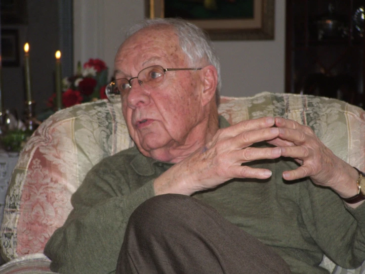 an old man in glasses sits on the sofa with his hands on his chest
