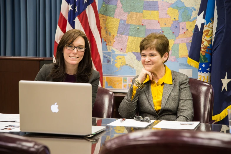 two women sitting in front of laptops next to a wall with maps