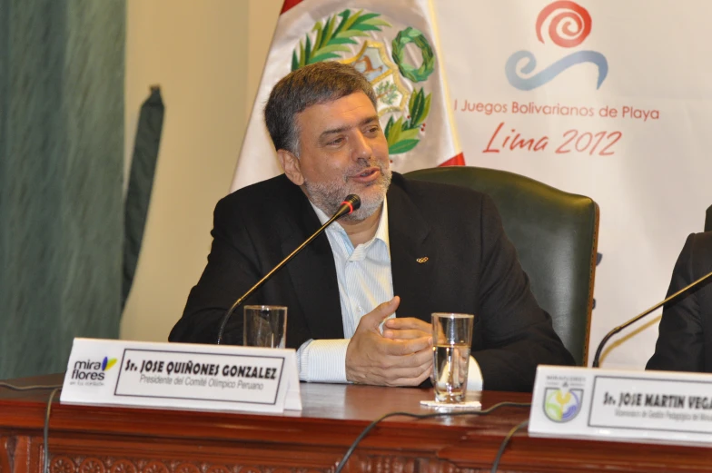 two men sitting in front of flags at a podium