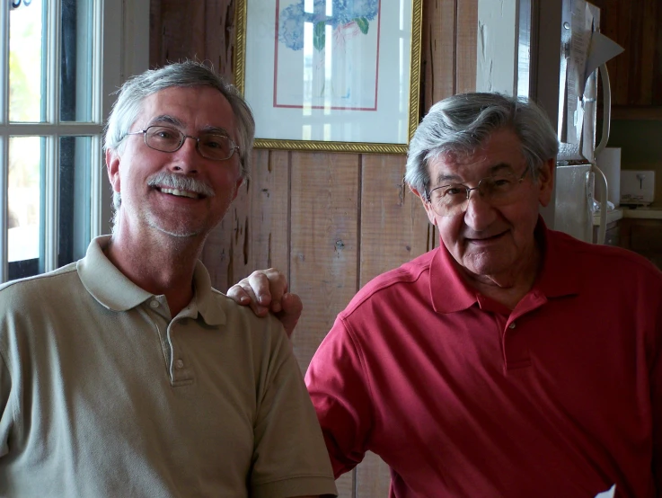 two older men standing next to each other in front of a wall with pictures on it