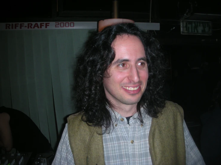 a man with black hair and a vest standing in front of a room