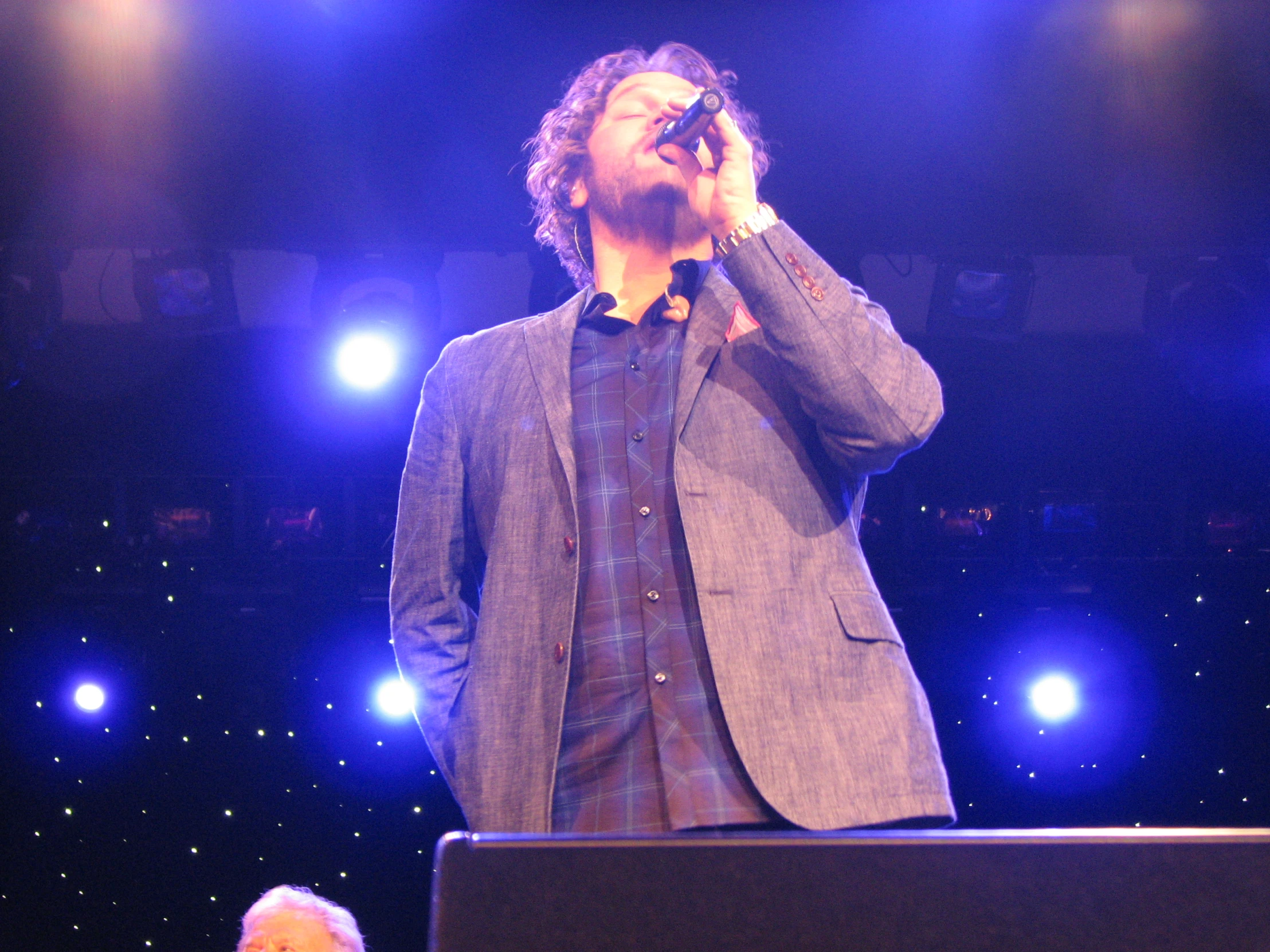 a man on stage wearing a gray jacket and blue shirt with his hands over his face