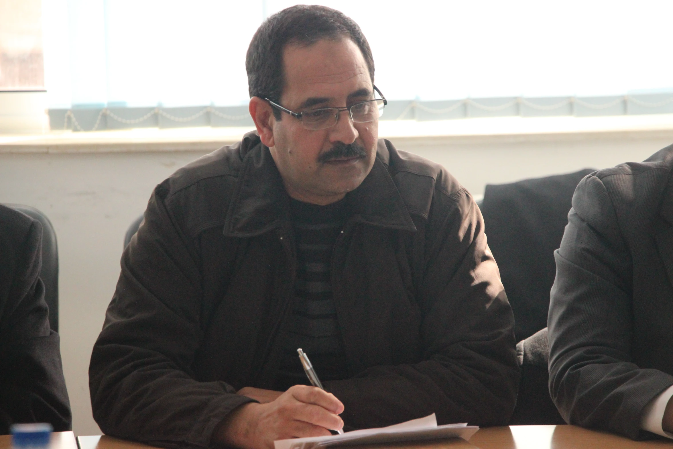 two men sitting at a table, one is holding a pen and writing