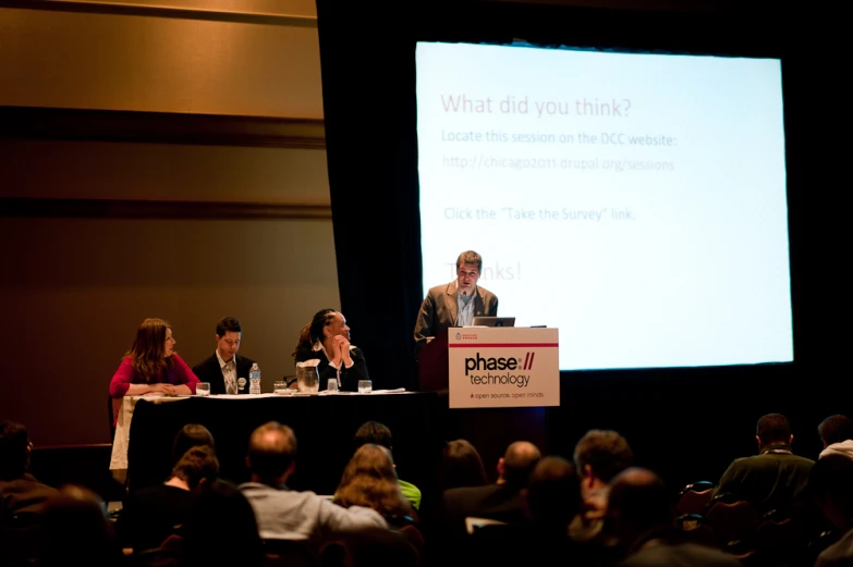 several people sitting at a table with microphones in front of a projection screen