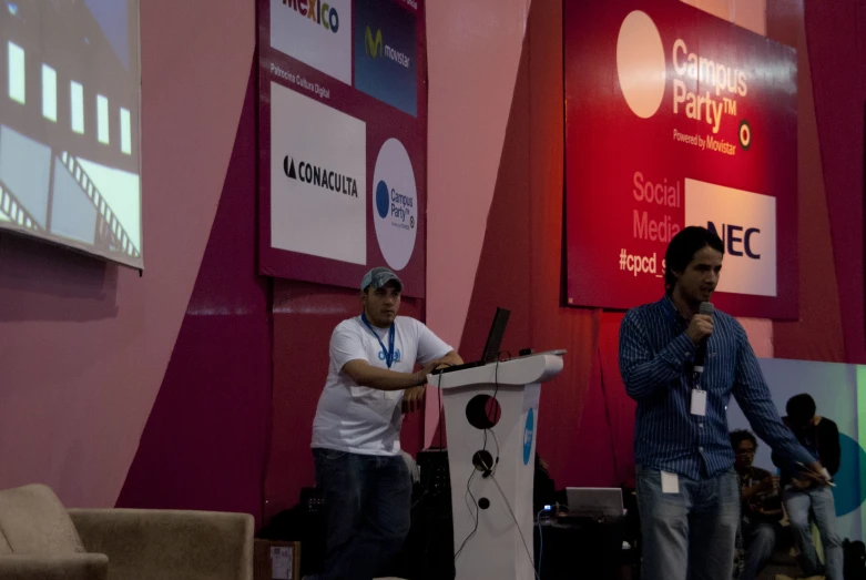 two men are speaking and holding microphones in front of a screen