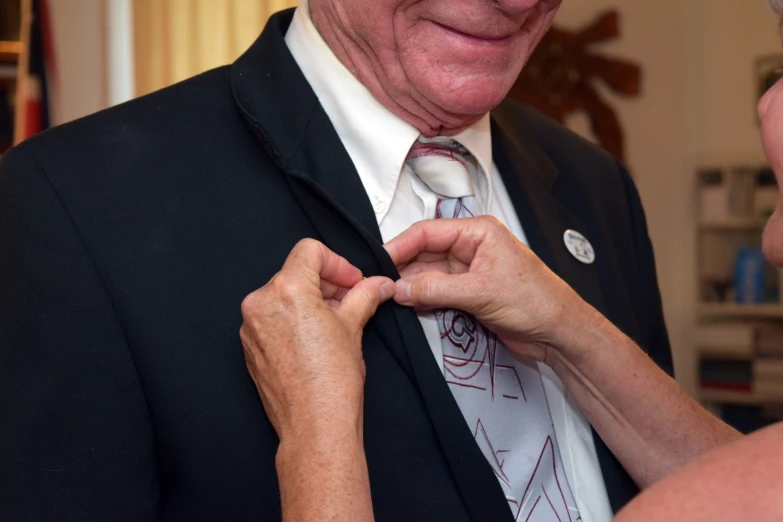 an old man with glasses adjusting a tie while standing next to an elderly woman