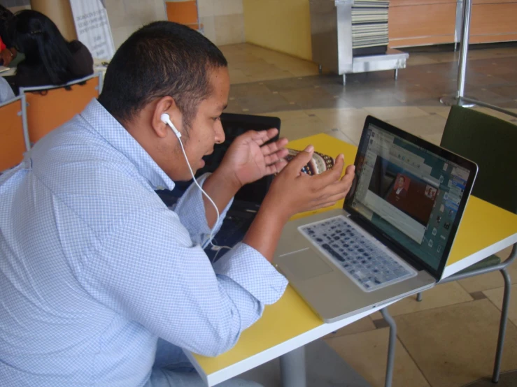 a man sits at a yellow table using his cell phone, in front of his laptop