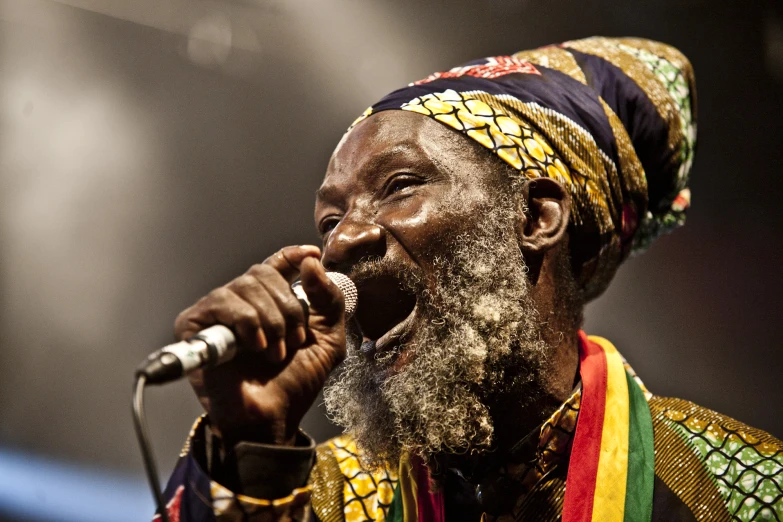 an african man wearing a brightly colored head scarf sings into a microphone