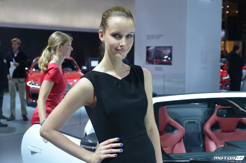 a lady in a black dress posing for the camera at a car show