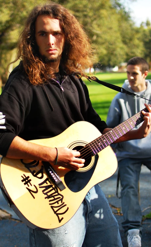 a young man with long hair plays an acoustic guitar outside