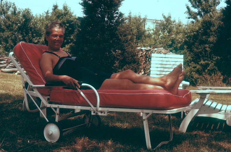 man on red lawn chaise lounge with trees and water behind him