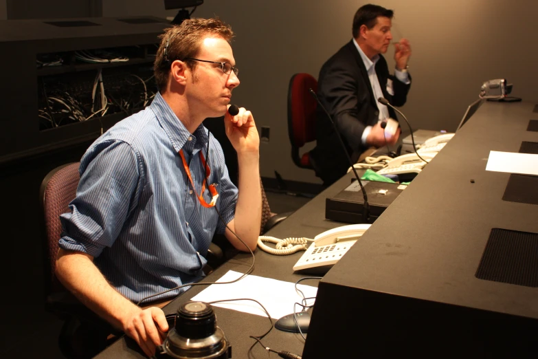 two guys at a desk one has a camera in his hand the other has an earplug