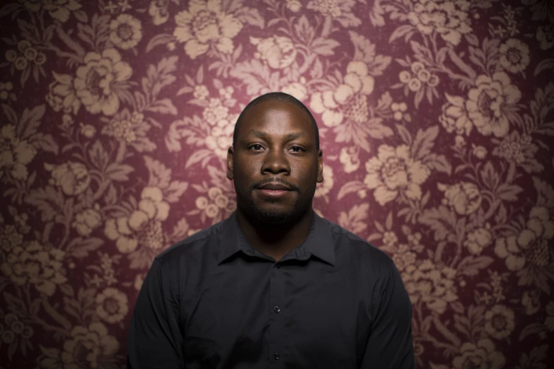 the man looks directly into the camera as he stands in front of a floral patterned wallpaper