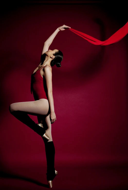 a woman in a red leotard stands against a red wall and holds a long ribbon